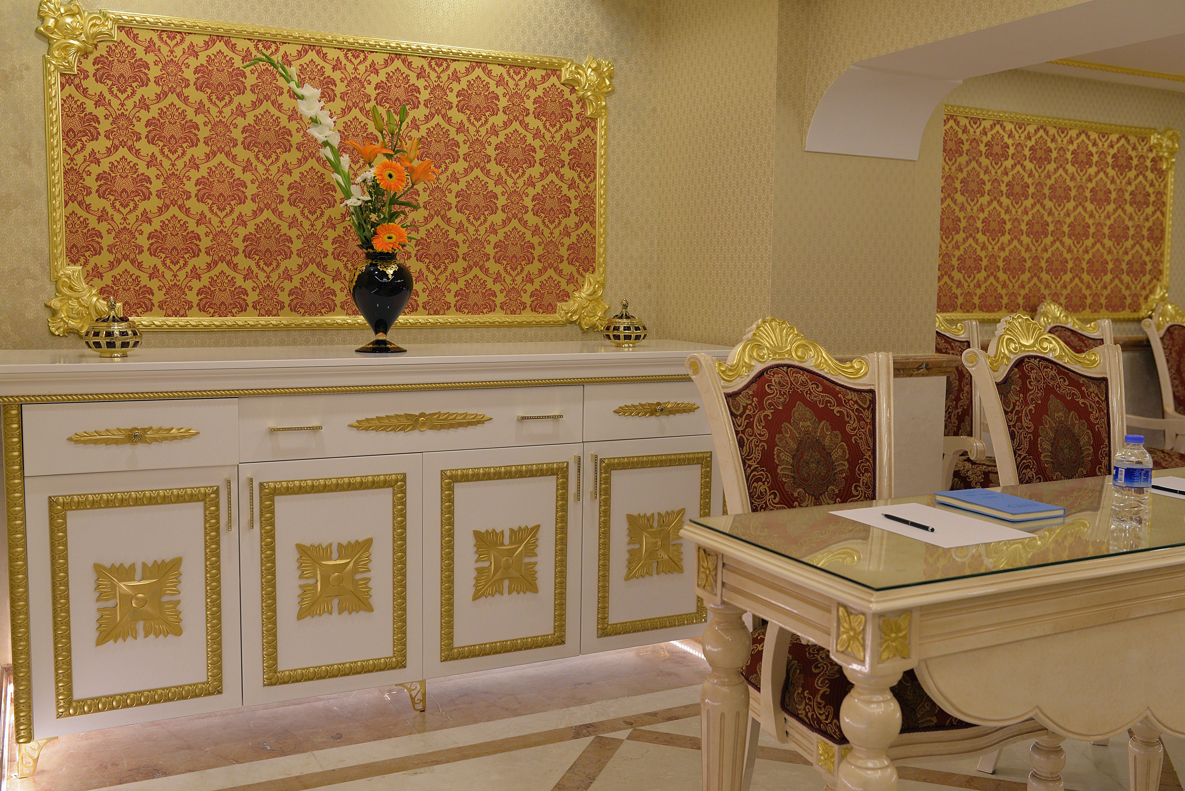 A room with a white and gold ornate cabinet, a glass table with a blue notebook and pen, and a vase with flowers.