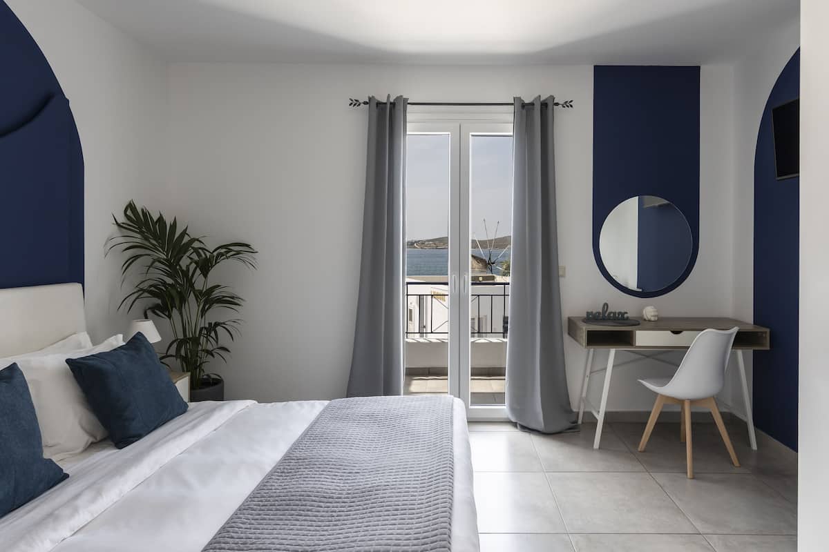 Un dormitorio moderno con cama, escritorio, silla, planta en maceta y vista al mar a través de un ventanal.