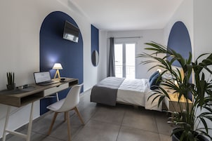 Un dormitorio con cama, escritorio con computadora portátil, silla, televisor montado en la pared y ventana con cortinas.