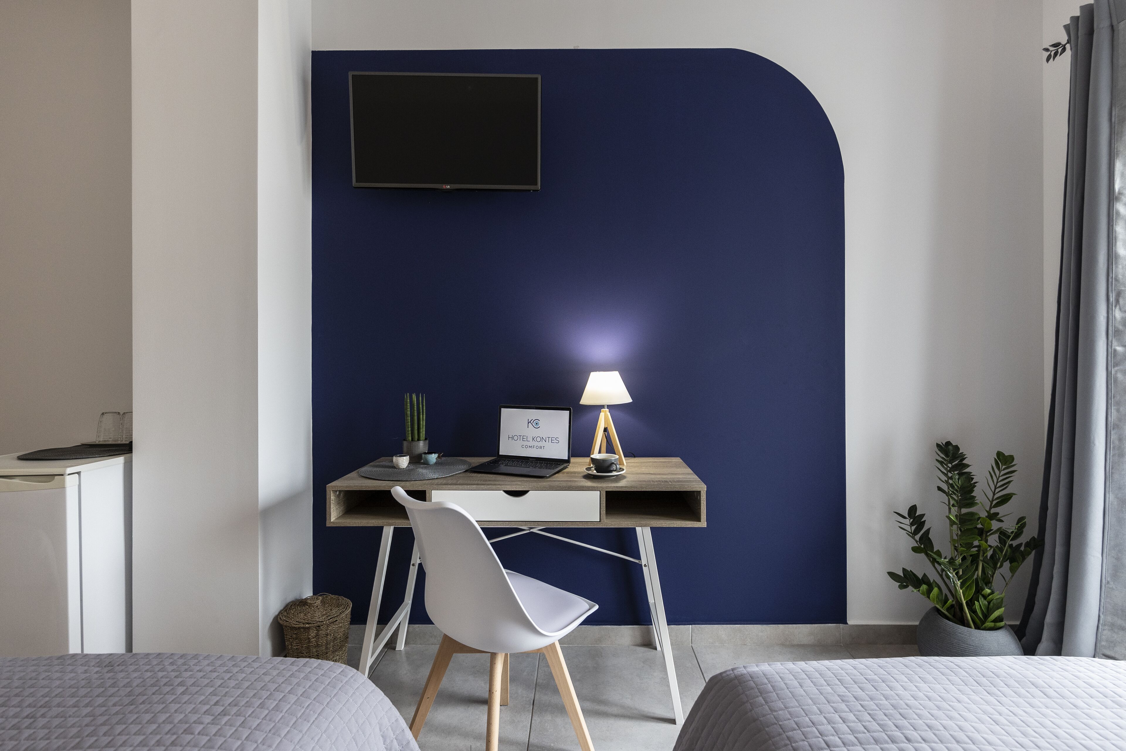 A hotel room with a desk, a laptop, a lamp, a potted plant, and two beds.