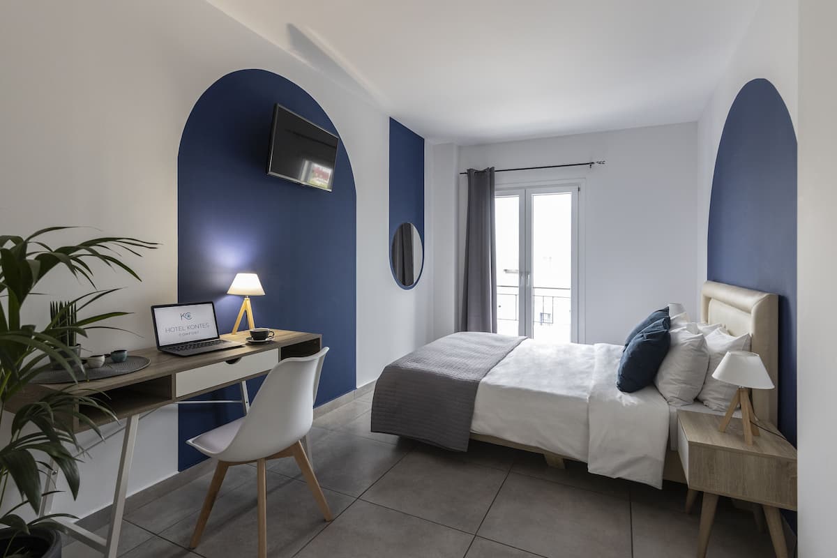 Un dormitorio con cama, mesita de noche, lámpara, escritorio con una computadora portátil, silla y una planta en maceta.