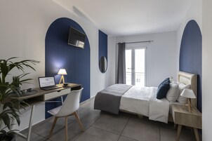Un dormitorio con cama, mesita de noche, lámpara, escritorio con una computadora portátil, silla y una planta en maceta.