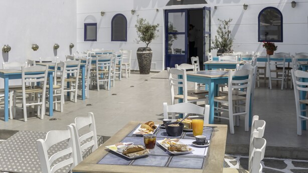 Uma área de refeições ao ar livre com mesas e cadeiras brancas e azuis, um buffet com comida e bebidas, e um edifício com a placa 'HOTEL KONTES'.
