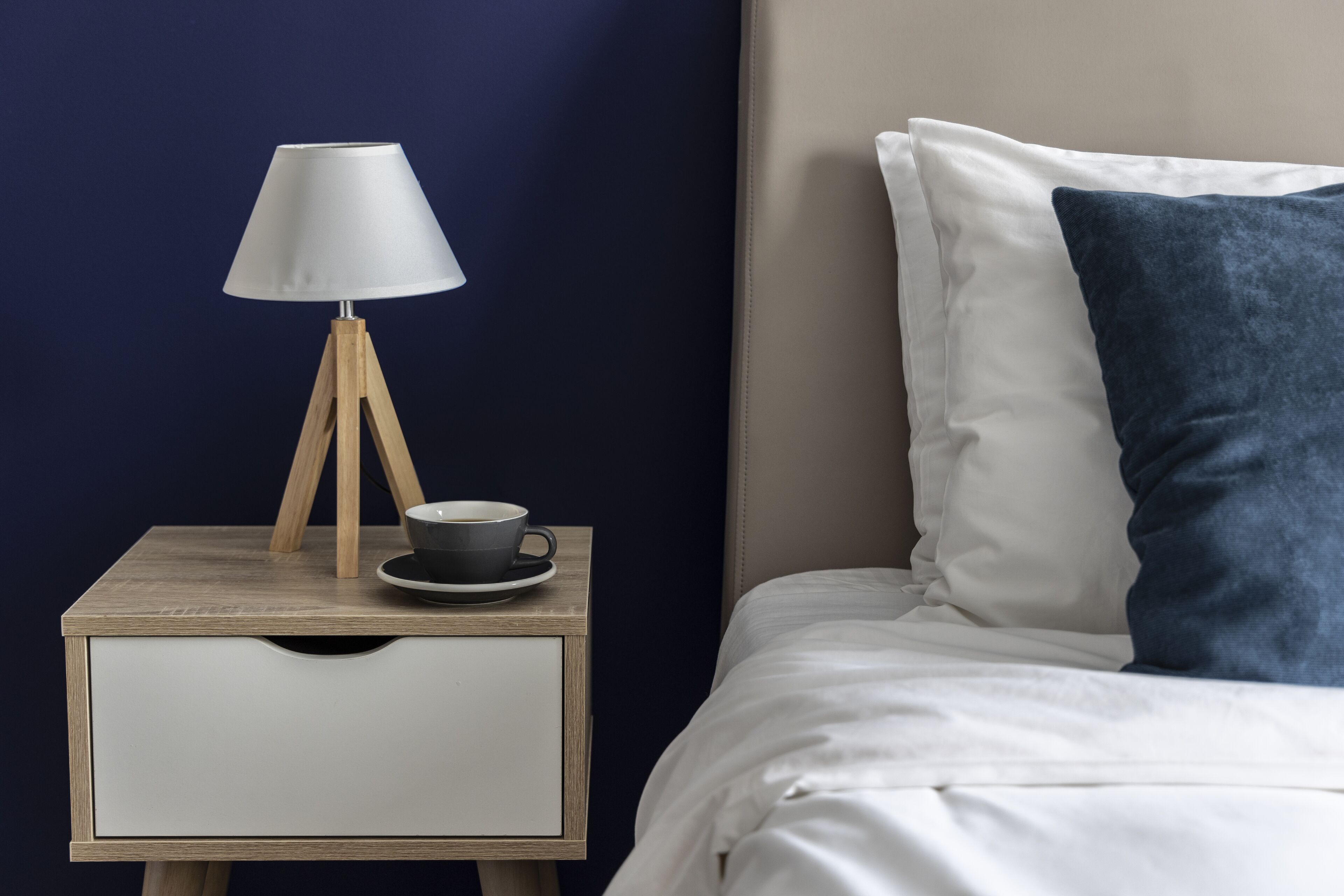 Ein ordentlich bezogenes Bett mit weißer Bettwäsche und einem blauen Kissen, ein Nachttisch mit einer Lampe und einer Tasse, sowie eine dunkelblaue Wand.