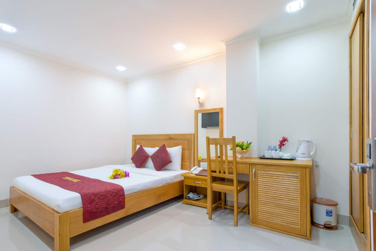 A hotel room with a wooden bed, a desk with a chair, a small trash bin, and a wall-mounted TV.