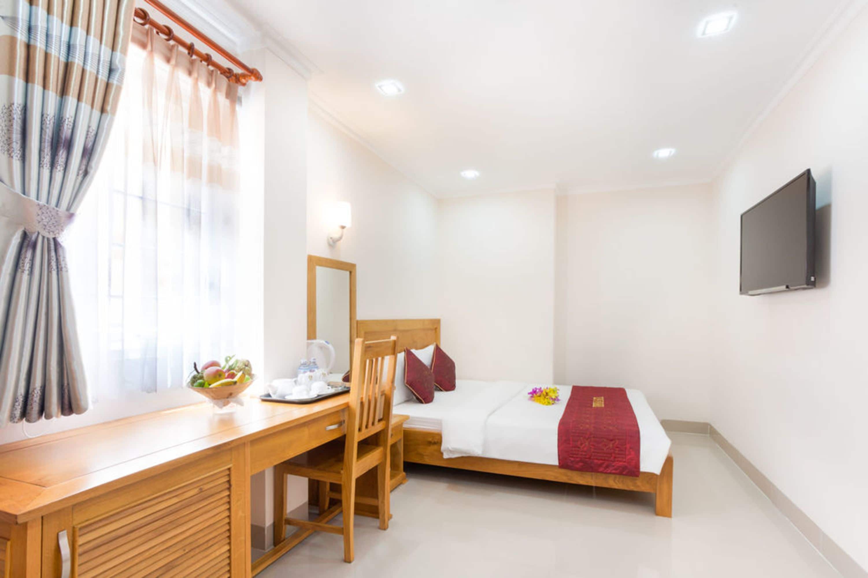 Hotelkamer met een houten bureau, een stoel, een bed met een rode deken, een nachtkastje met een schaal fruit, en een wandtelevisie.