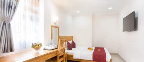 A hotel room with a wooden desk, a chair, a bed with a red blanket, a bedside table with a fruit bowl, and a mounted TV.