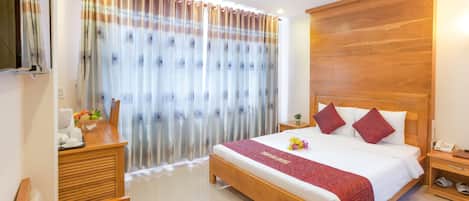 A hotel room with a wooden bed, a wooden nightstand, a wooden desk, a chair, and a TV mounted on the wall.