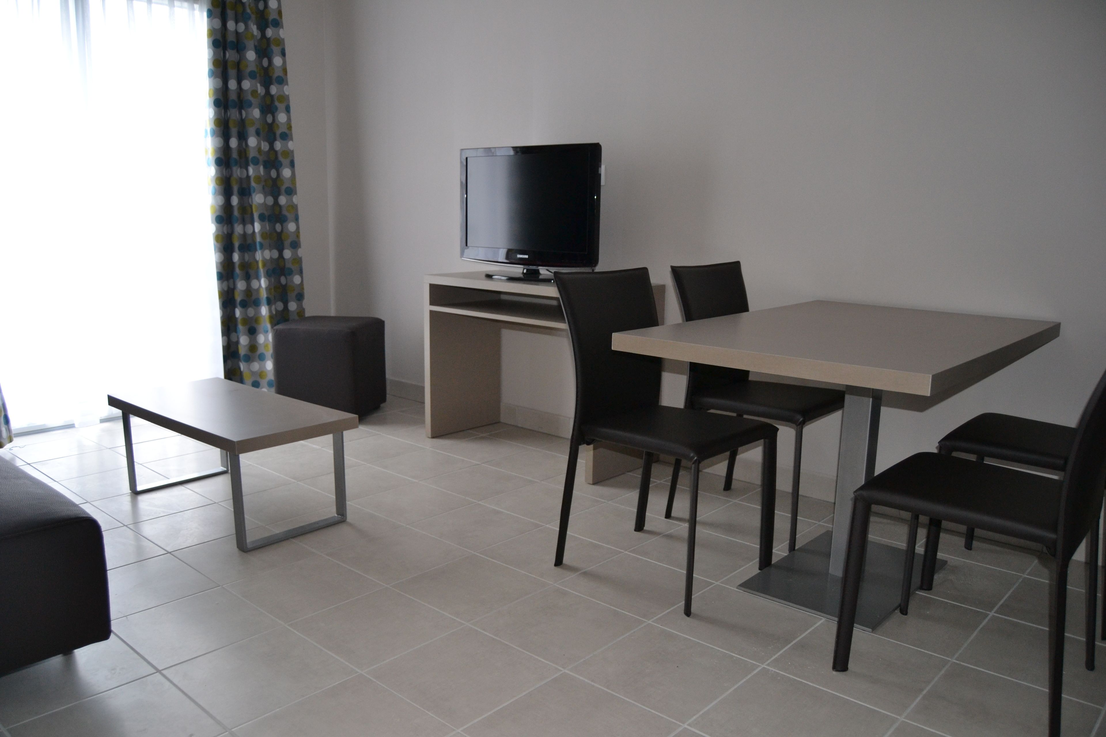 A modern living room with a flat-screen TV on a stand, a dining table with four chairs, a sofa, and a coffee table.