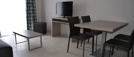A modern living room with a flat-screen TV on a stand, a dining table with four chairs, a sofa, and a coffee table.