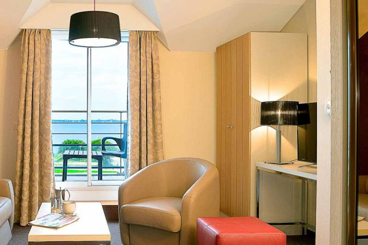 Habitación de hotel con sofá, sillón, una mesita y televisión. Se ve un ventanal con cortinas y vistas al exterior con vegetación.
