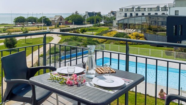 En balkon med et bord dækket til to, med udsigt over en swimmingpool og en anlagt have.