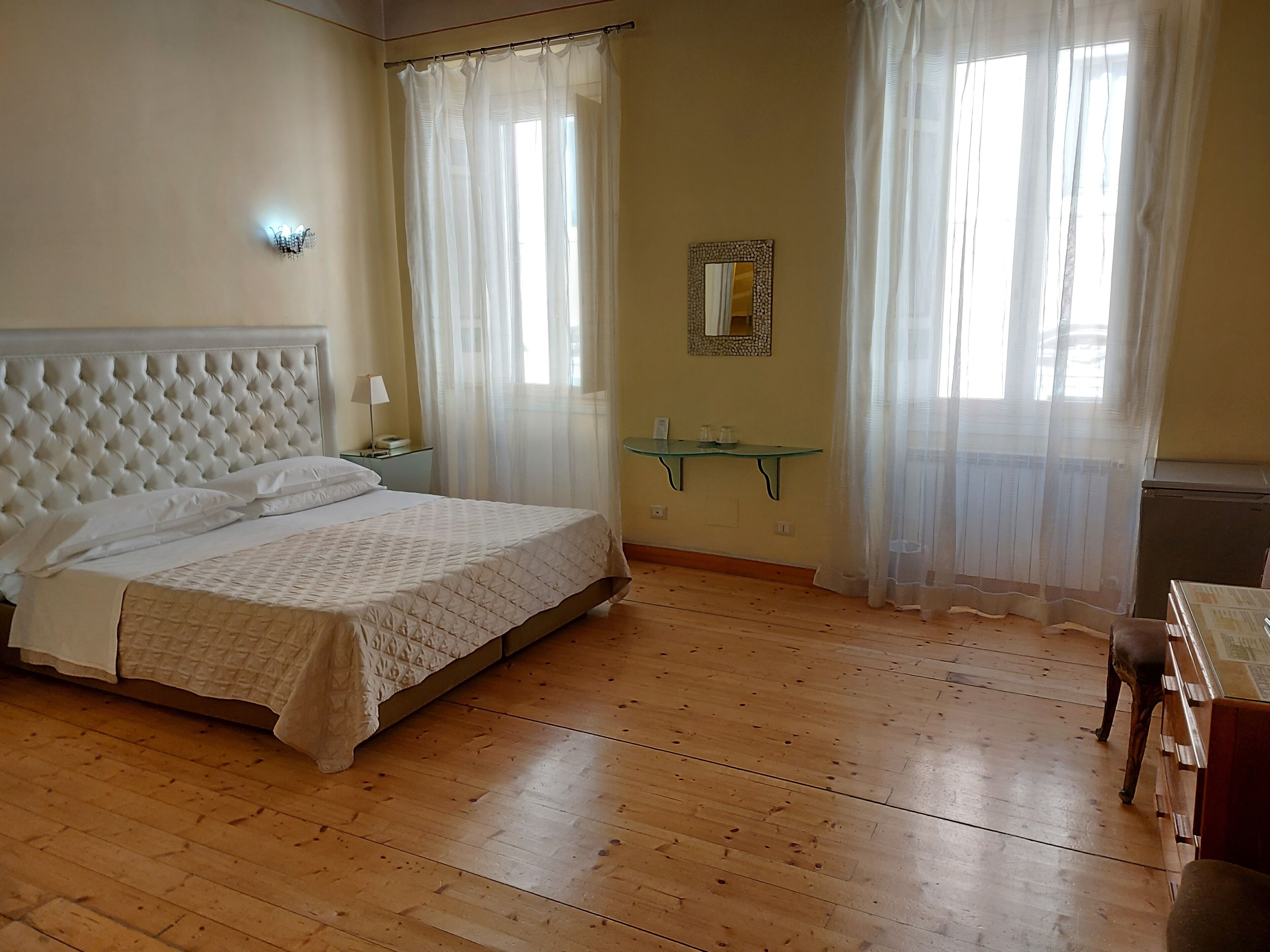 Ein Schlafzimmer mit einer gepolsterten Kopfteil, einem Bett mit gesteppter Bettdecke, einem Nachttisch mit Lampe, einem Spiegel an der Wand und einem kleinen Tisch mit Glasplatte.
