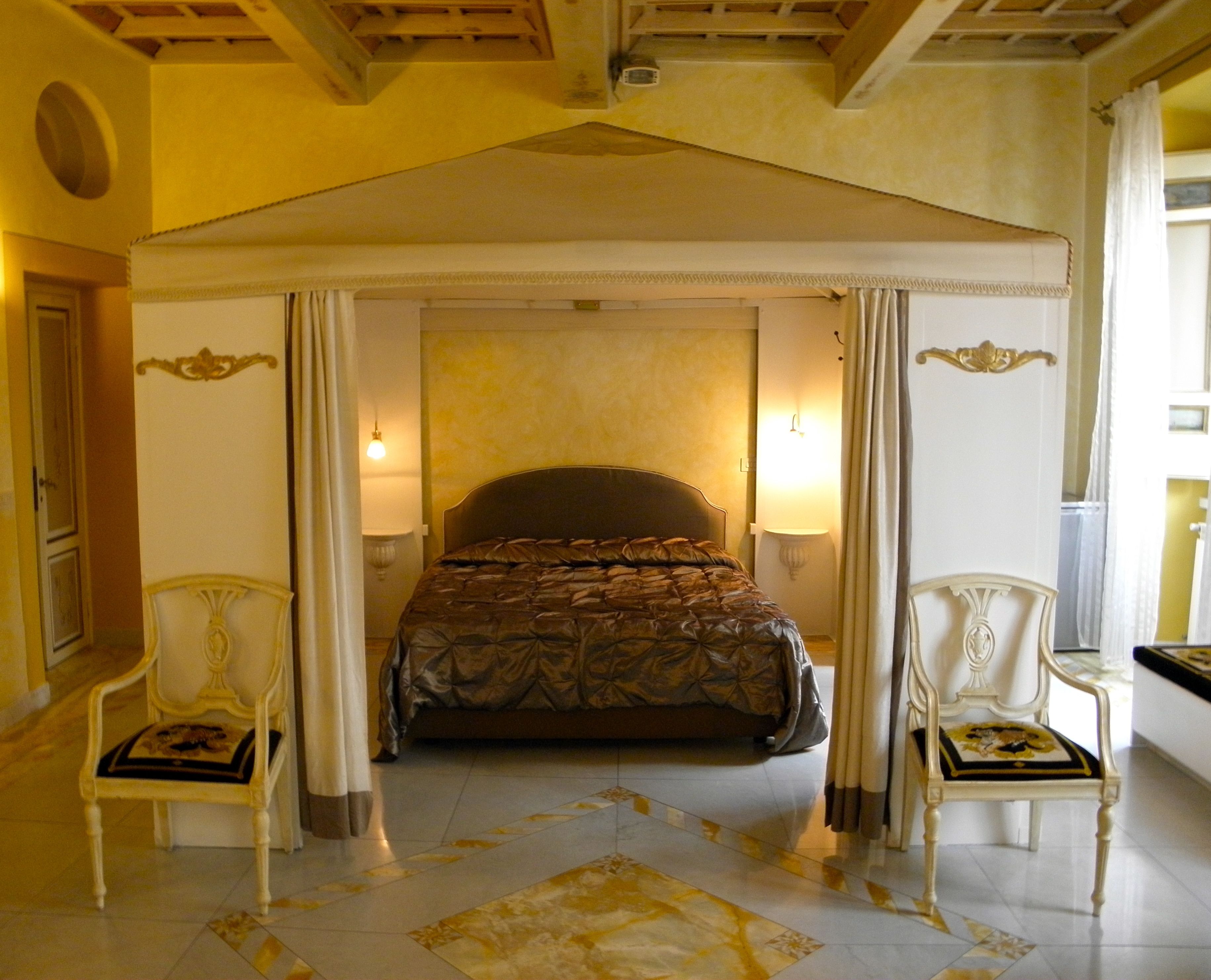 Una cama con dosel con un edredón estampado oscuro, flanqueada por dos sillas ornamentales con cojines negros y dorados.