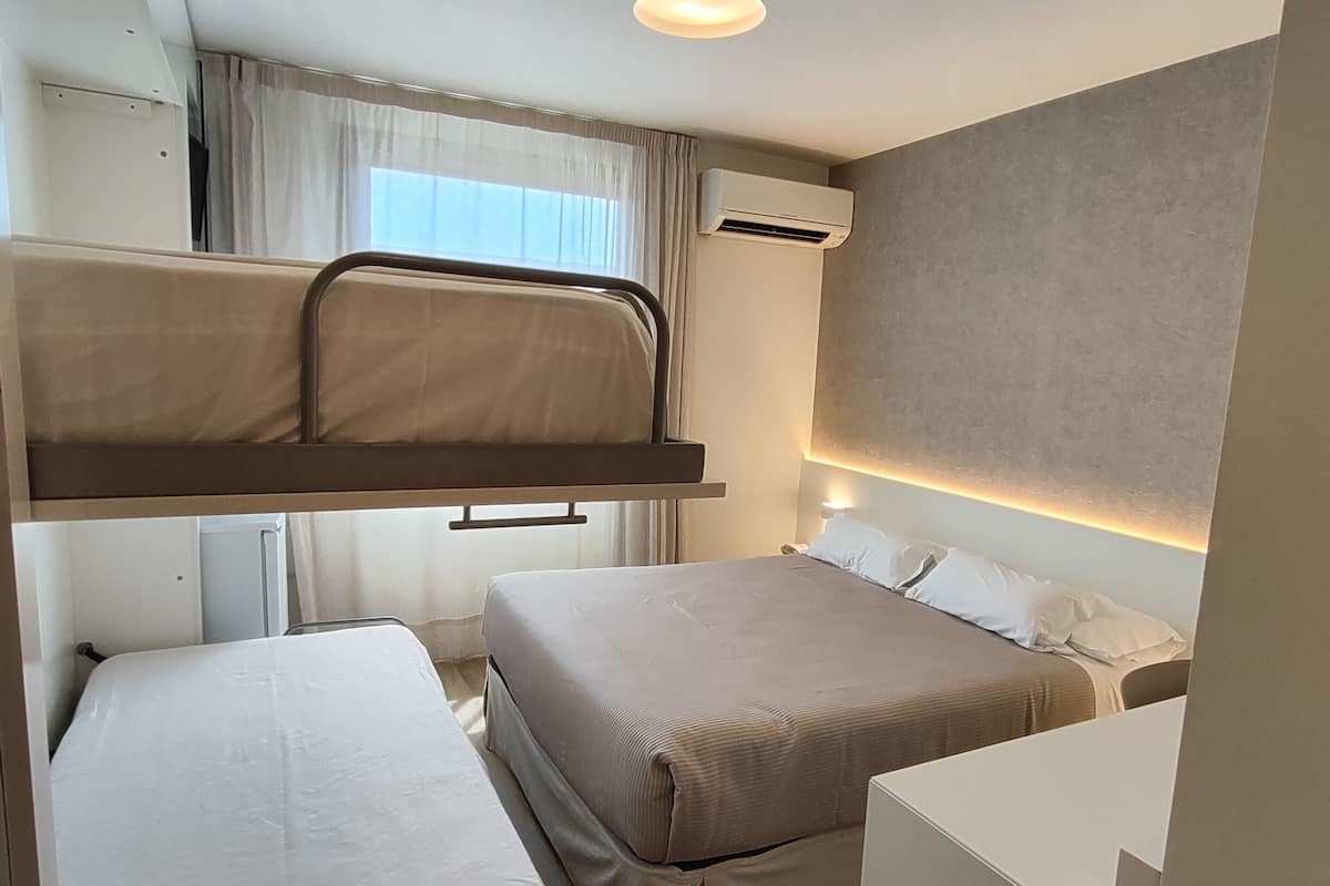 Quarto de hotel compacto com beliches, uma escrivaninha e uma pequena cozinha equipada.
