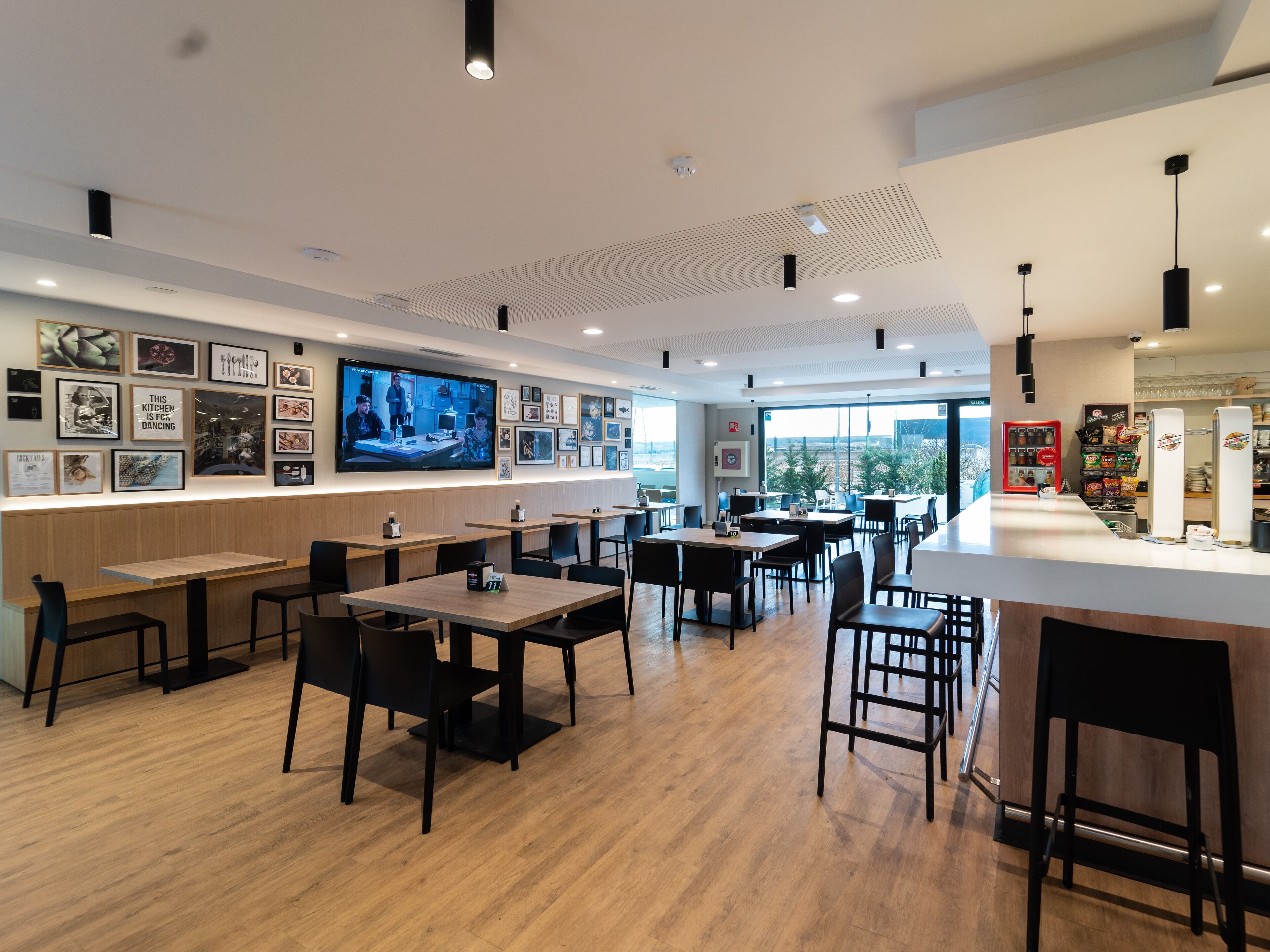 A modern restaurant interior with wooden tables and chairs, a large TV screen on the wall, and a vending machine.