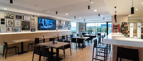 A modern restaurant interior with wooden tables and chairs, a large TV screen on the wall, and a vending machine.