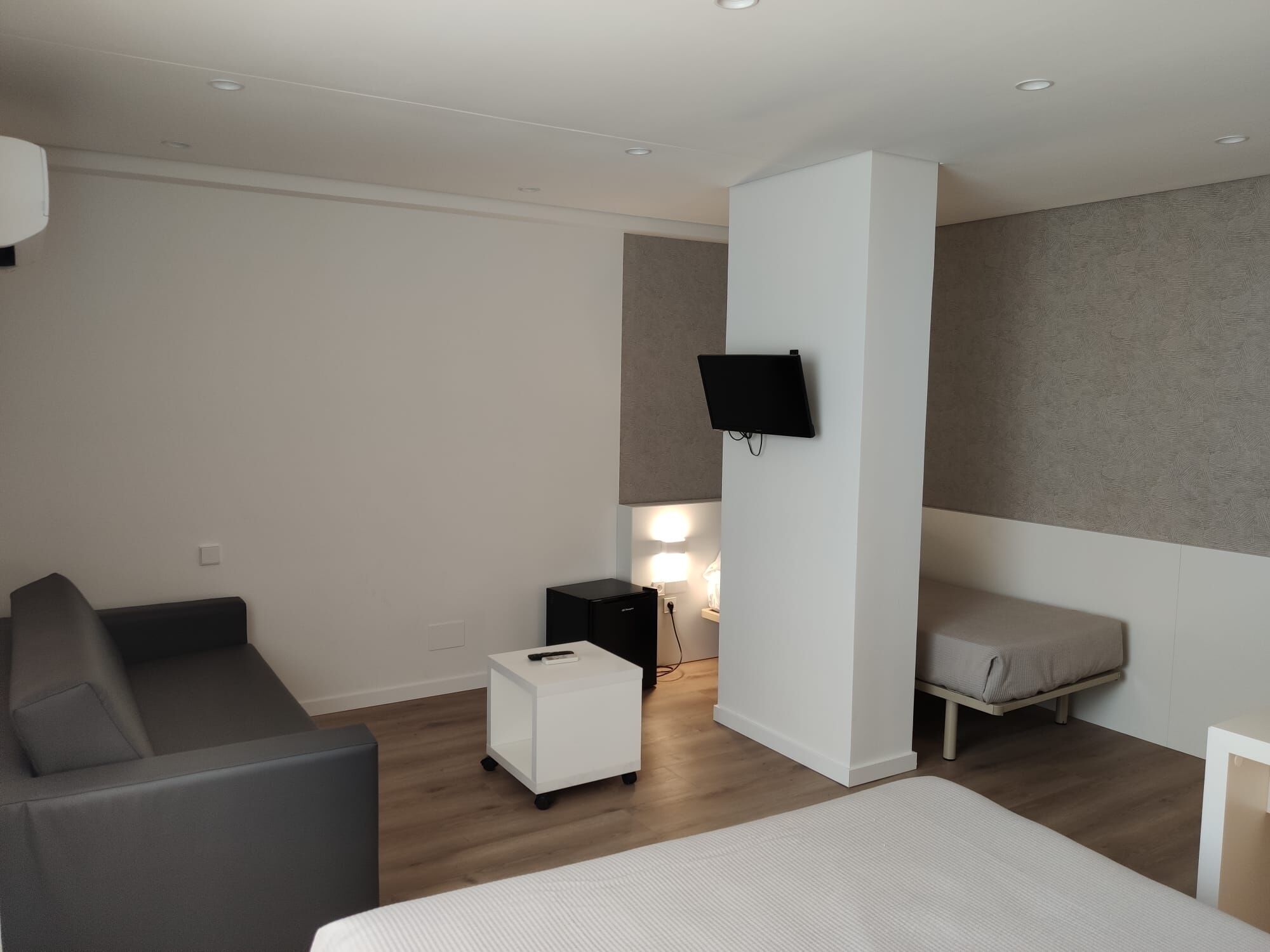 Una habitación de hotel moderna con una cama, un sofá, una mesita blanca y un televisor montado en la pared, además de una lámpara.