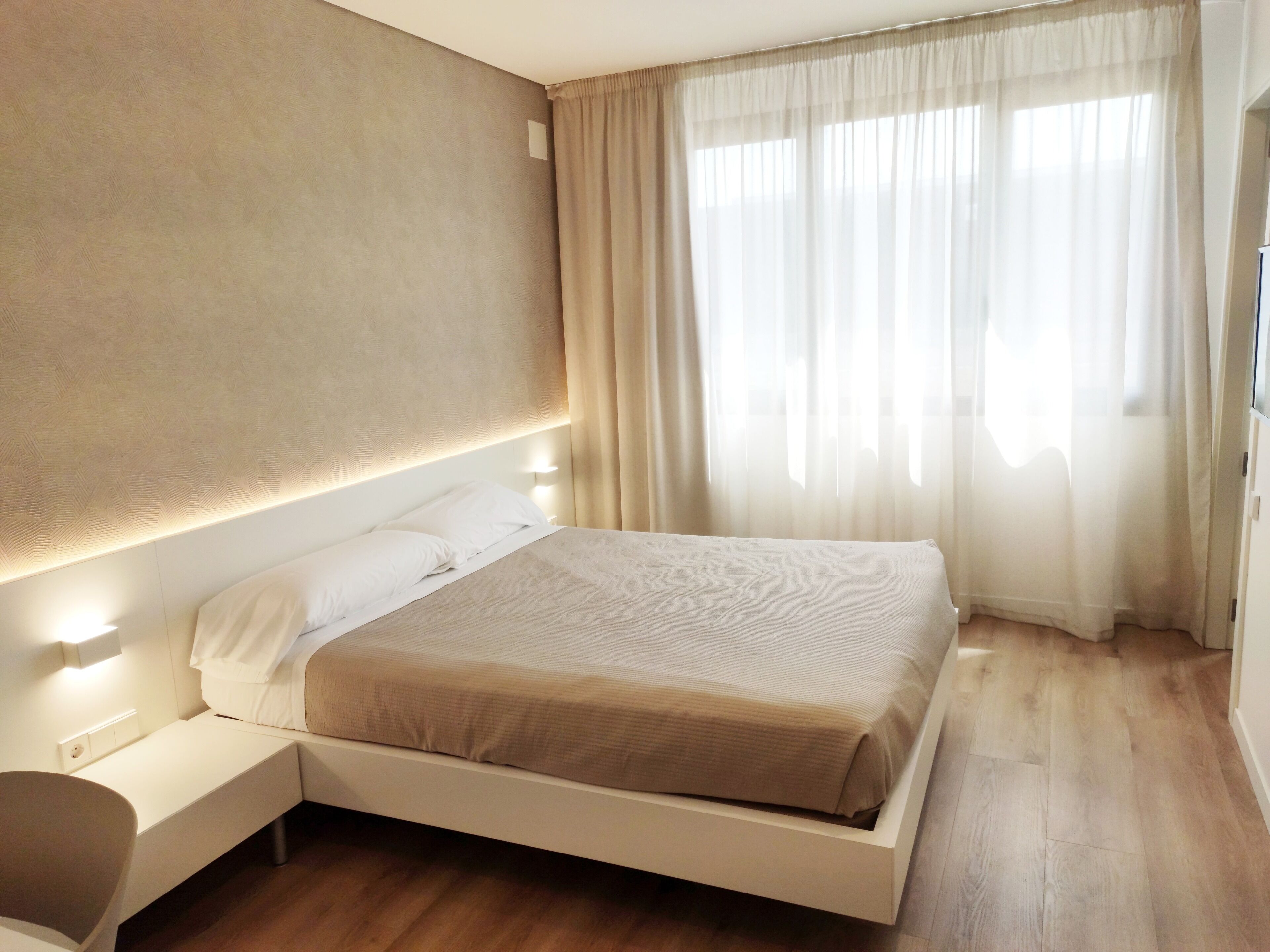 Un dormitorio moderno con una cama, mesitas de noche y un ventanal con cortinas translúcidas.