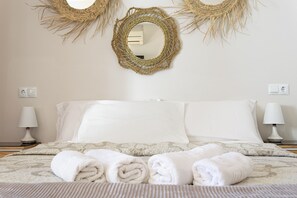 A neatly made bed with rolled towels, two bedside lamps, and two wall-mounted woven mirrors.