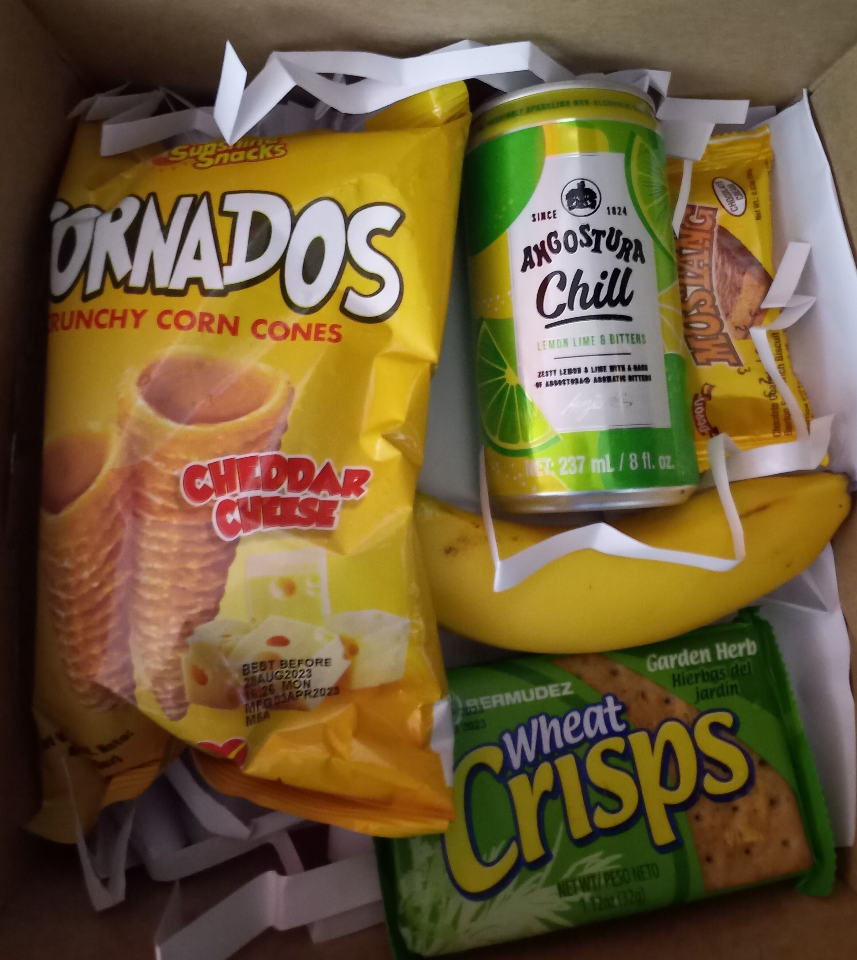 A box containing snacks and drinks including corn snacks, a can of Angostura Chill, and wheat crisps.