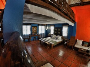 A room with a bed, a desk, and a chair. There are windows with curtains and a painting on the wall.