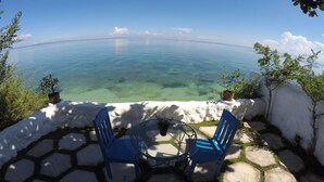 Une terrasse avec deux chaises bleues et une table, d’où l’on peut admirer une mer bleue et limpide, avec une côte lointaine à l’horizon.