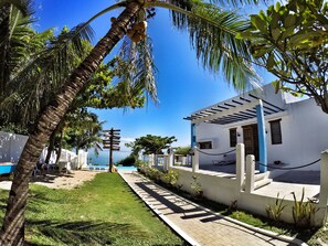 A white building with a covered patio, a clear blue sky, and a palm tree with coconuts.