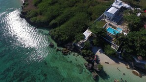 Vue aérienne d’une propriété située sur la côte, avec une piscine, une végétation luxuriante et une eau turquoise cristalline.
