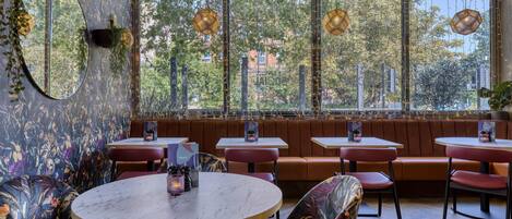 A restaurant with round tables, cushioned seating, and large windows with a view of trees and buildings.