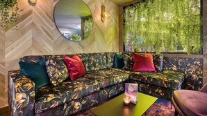 A modern living room with a herringbone-patterned wall, a round mirror, a sofa with colorful cushions, and a window with green foliage. - Premier Inn London Hampstead (London)