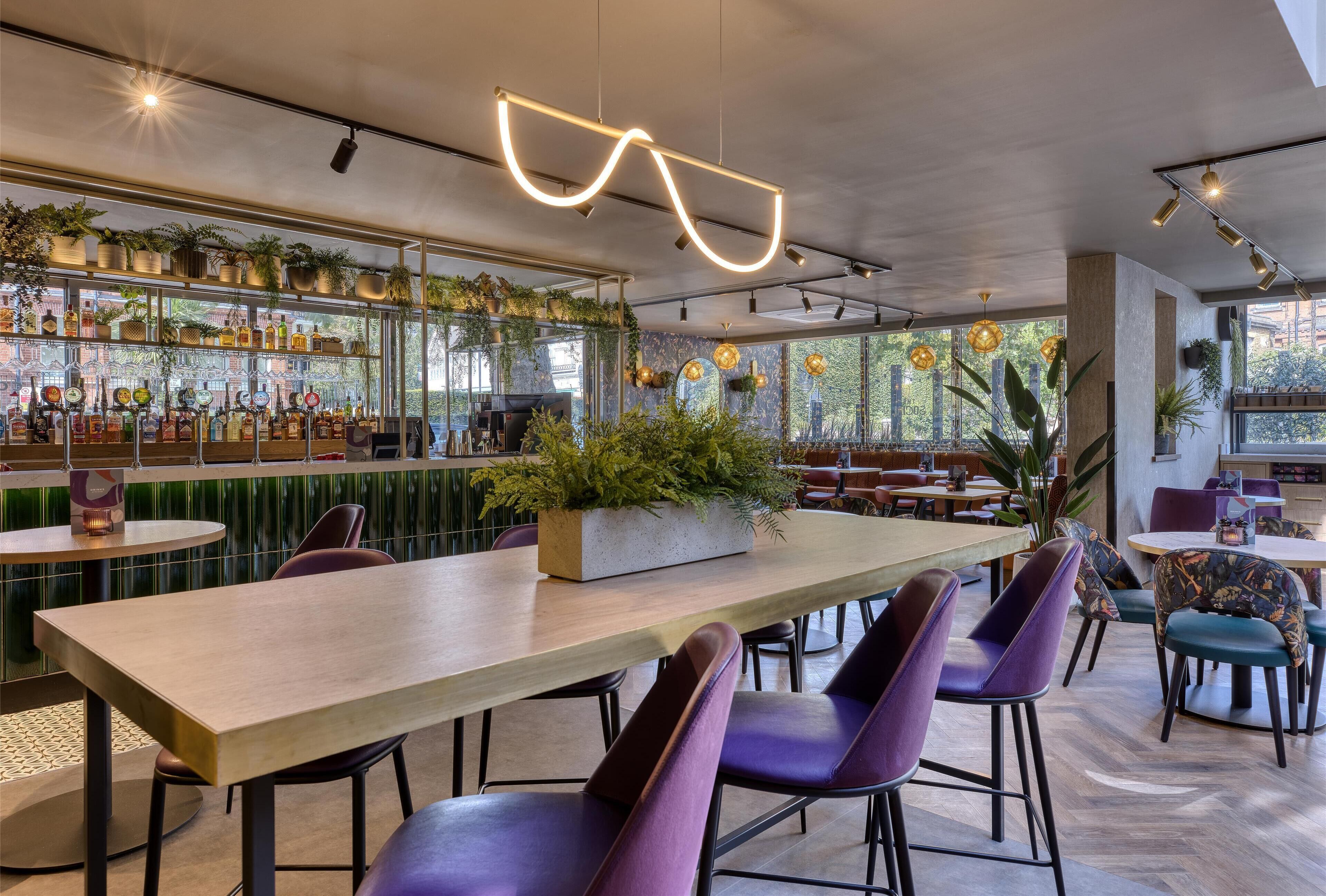A modern bar area with seating, a long table, and a variety of plants and bottles.