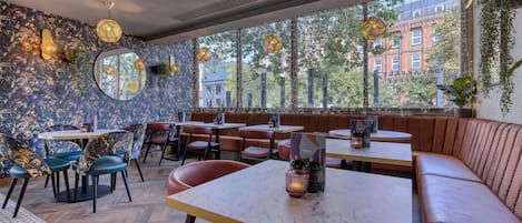 Un interior de restaurante con mesas redondas, asientos acolchados, ventanas amplias e iluminación decorativa.