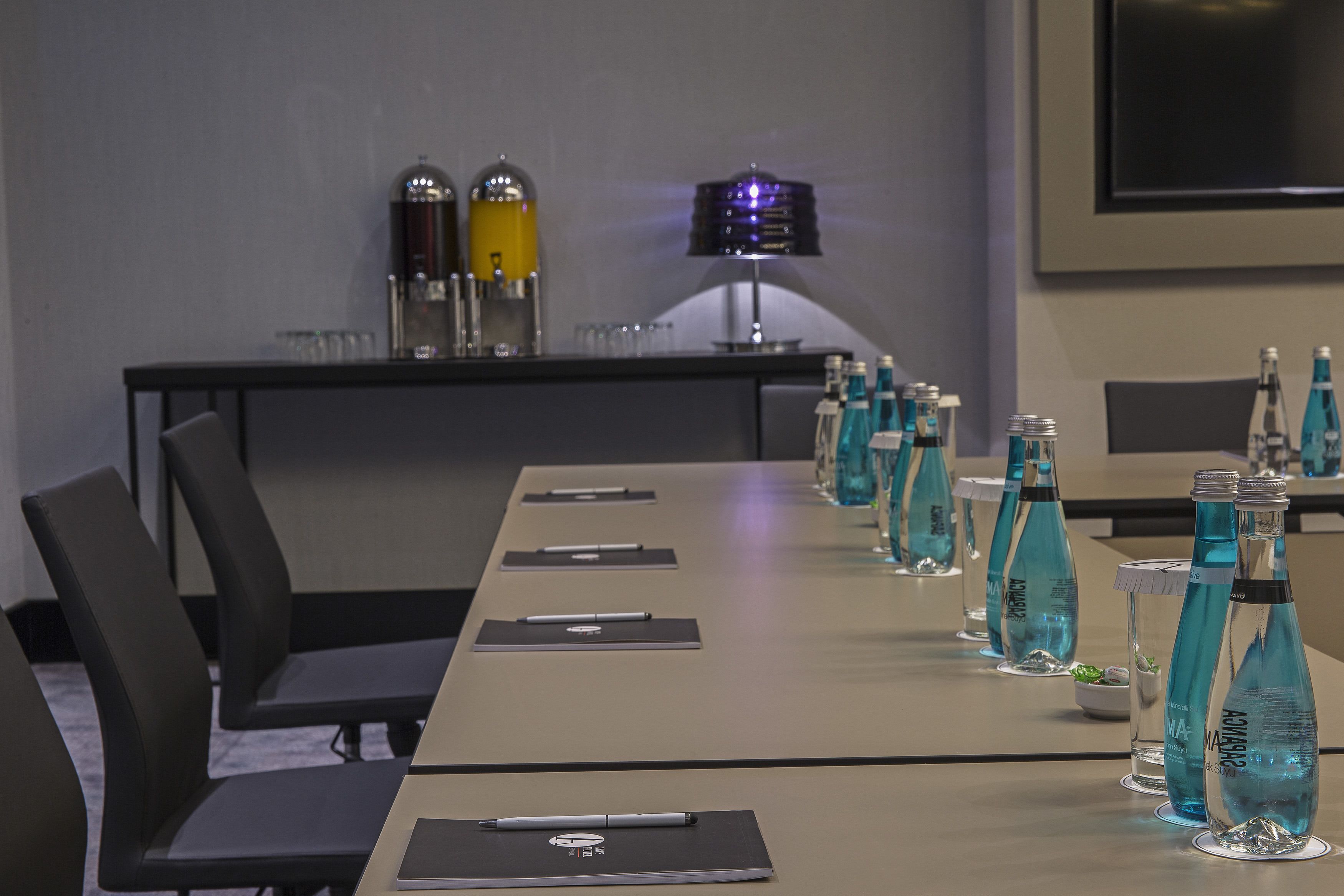 Un salón de conferencias con una larga mesa, sillas, botellas de agua y un proyector.