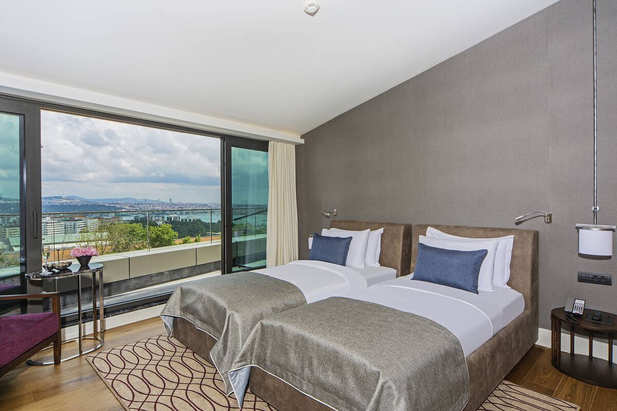 A modern hotel room with two beds, a large window with a city view, a small table with a vase, and a chair.