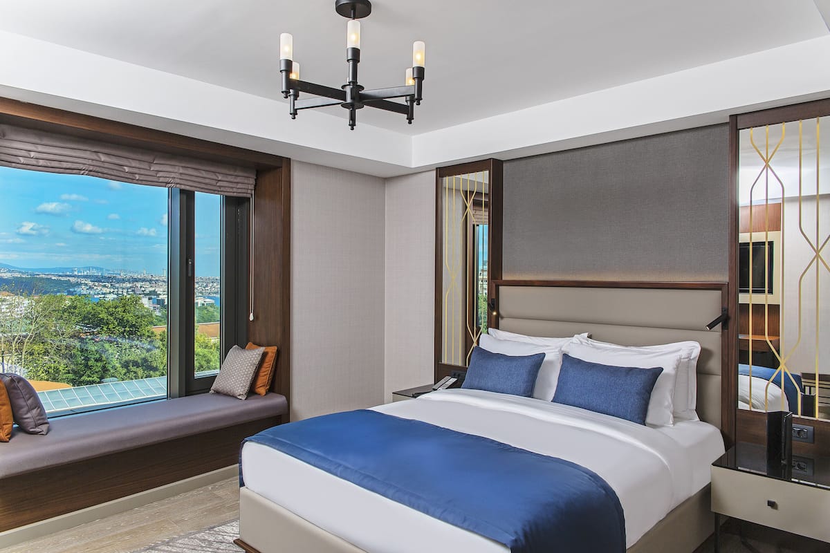 A modern hotel room with a large bed, a view of the city, and a seating area by the window.