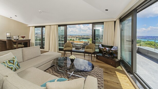 A modern living room with a sofa, chairs, a coffee table, and a TV. Large windows offer a city view. - Arts Hotel Istanbul - Special Class (Istanbul)