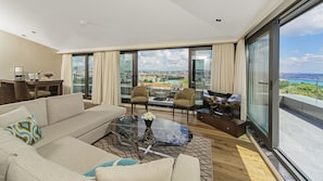 A modern living room with a sofa, chairs, a coffee table, and a TV. Large windows offer a city view.