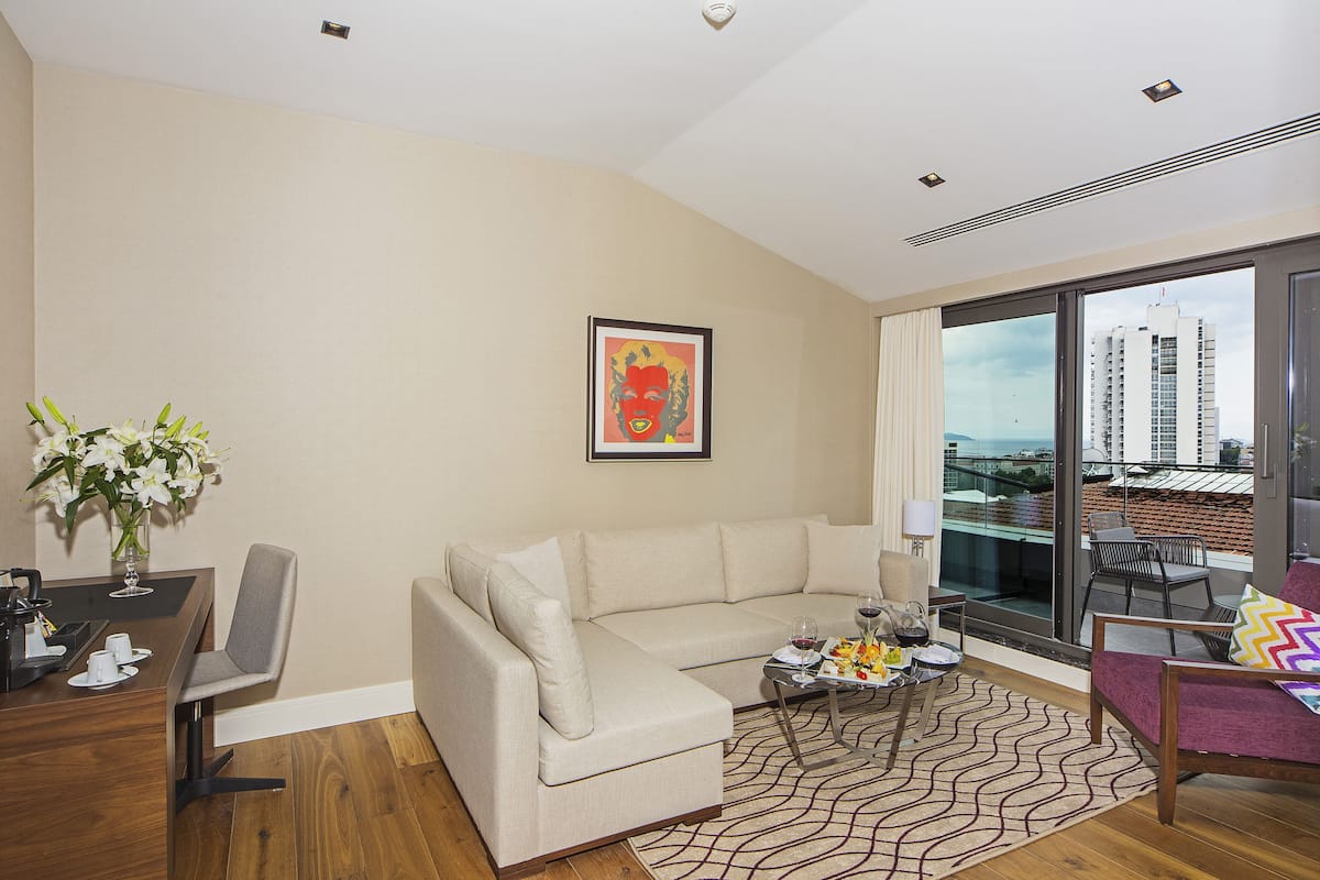A modern living room with a beige sofa, a dining table with chairs, a coffee table with drinks, and a large window with a city view.
