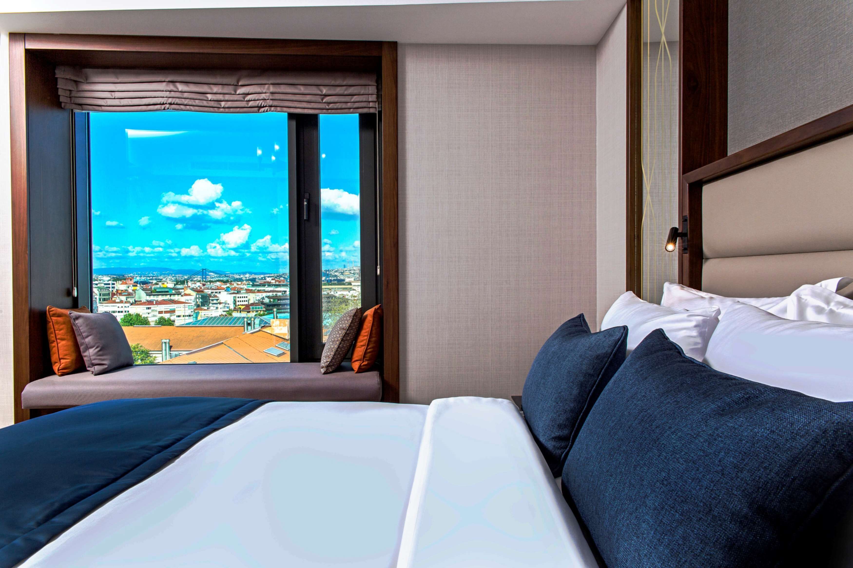 Habitació d'hotel amb un llit gran, vistes a la ciutat a través d'una finestra i una zona de seients amb coixins.