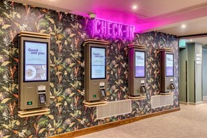 A check-in area with a 'CHECK-IN' sign above three self-service kiosks against a floral wallpaper backdrop. - Sleep Inn Tewkesbury (Malmesbury)
