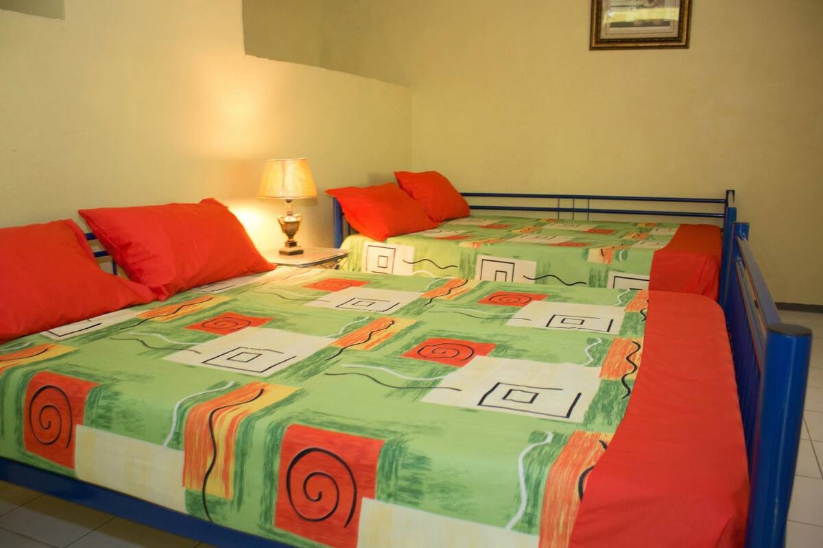 A room with two beds, each with red pillows and a colorful bedspread. There is a lamp on a bedside table.