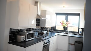 A modern kitchen with white cabinets, a black countertop, a gas stove, a microwave, and a window with a flower vase. - Canary Heights Apartments (London)