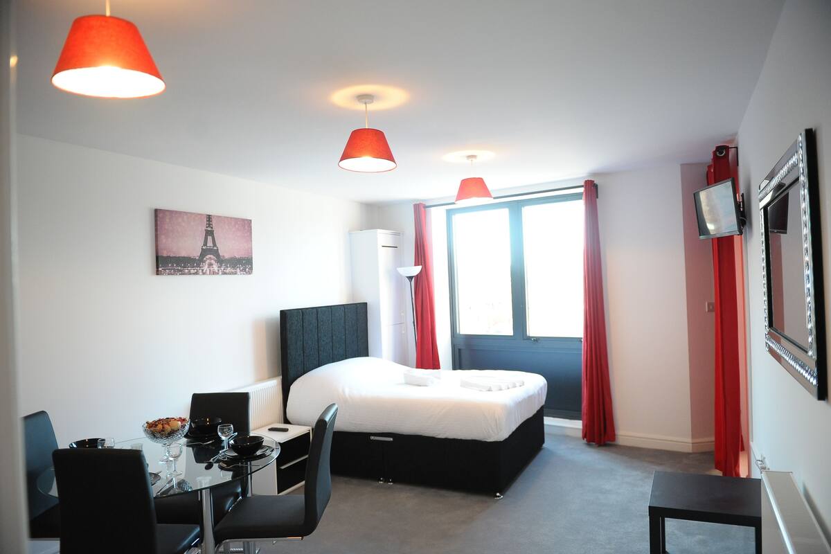 A modern hotel room with a bed, a desk, and a chair. There is a window with red curtains and a picture of the Eiffel Tower on the wall.