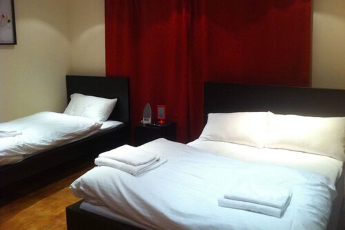 A hotel room with two single beds, each with white linens and pillows, a nightstand with a red lamp, and red curtains.