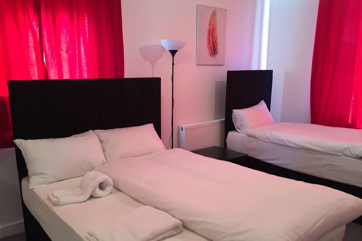A hotel room with two beds, red curtains, a floor lamp, and a painting on the wall.