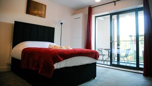 A bedroom with a large bed, a red blanket, a headboard, a floor lamp, and a sliding glass door leading to an outdoor area. - Canary Heights Apartments (London)