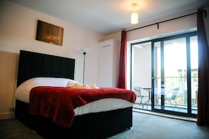 A bedroom with a large bed, a red blanket, a headboard, a floor lamp, and a sliding glass door leading to an outdoor area. - Canary Heights Apartments (London)