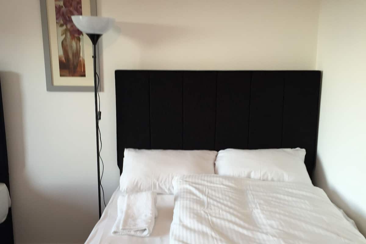 A neatly made bed with white linens, a black headboard, and a framed floral painting on the wall.