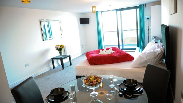 A hotel room with a bed, a dining table set for two, a TV mounted on the wall, and a window with curtains. - Canary Heights Apartments (London)